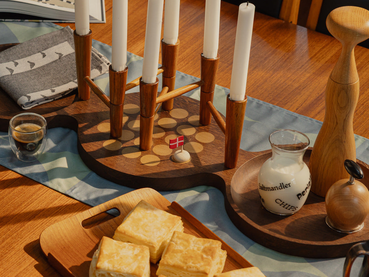 A round teak dining table set for a Danish jul gathering, with a wooden multi-arm candleholder, patterned napkins, and a small Danish flag.