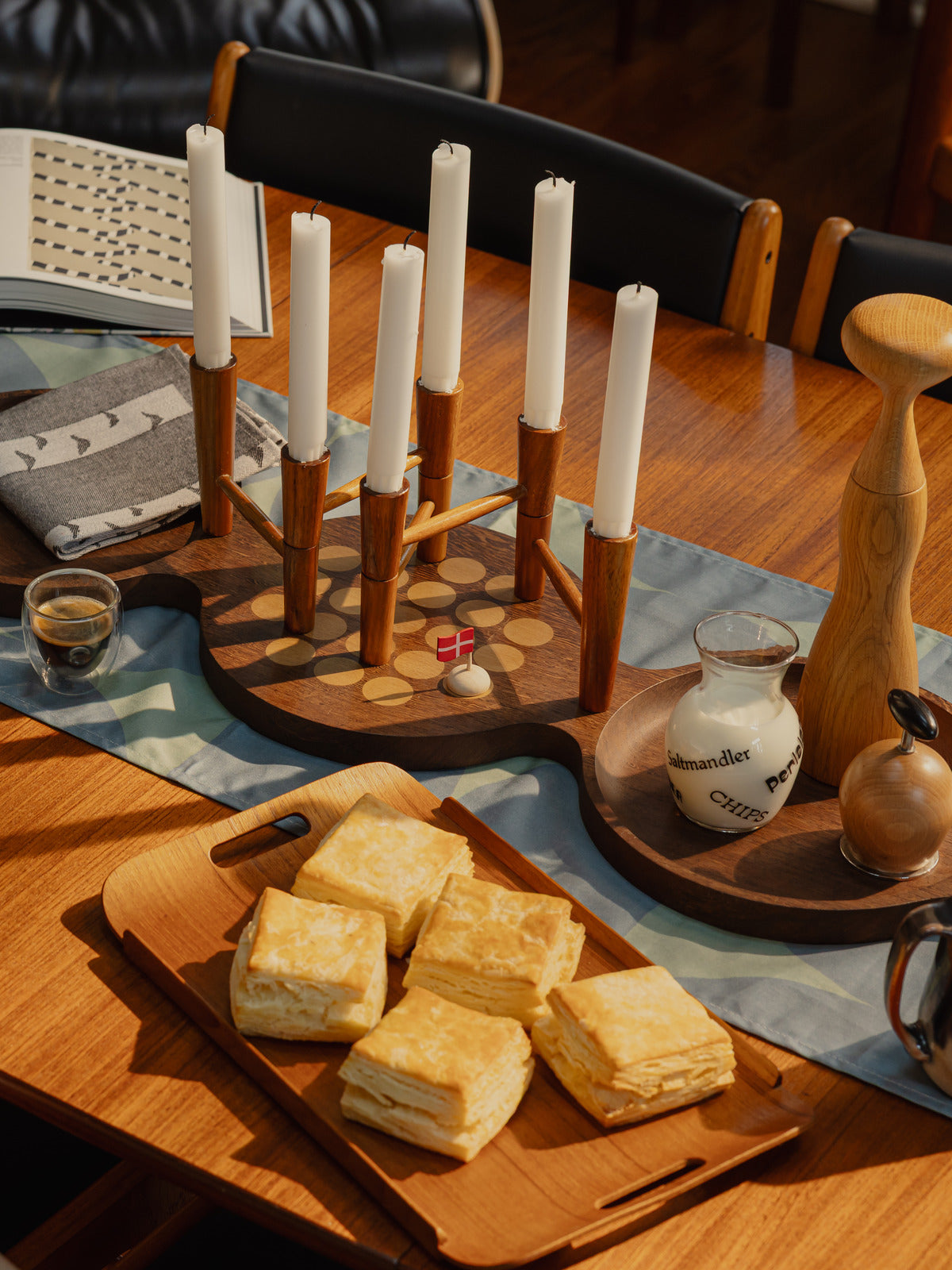 A round teak dining table set for a Danish jul gathering, with a wooden multi-arm candleholder, patterned napkins, and a small Danish flag.