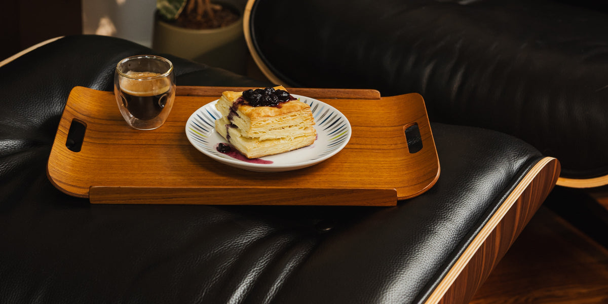 A teak tray with espresso and a pastry resting on a leather ottoman
