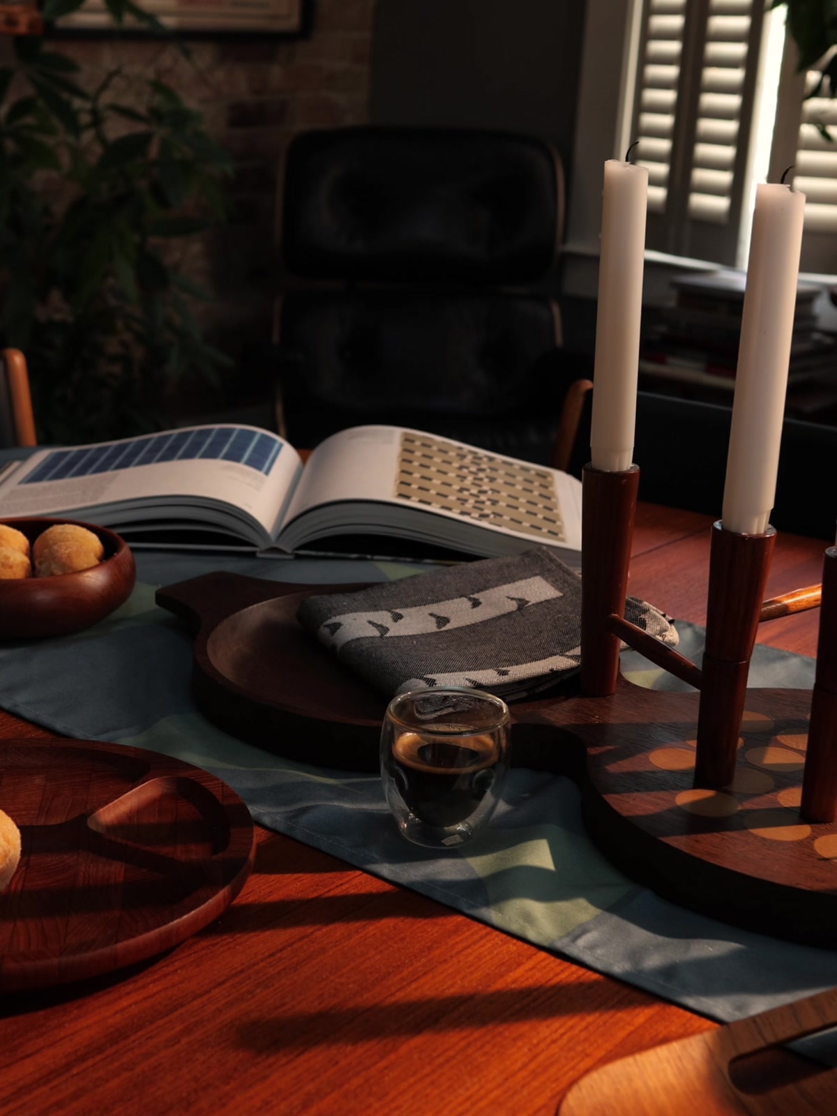 A dining table laid for morning coffee, with an open textile book, buns in a teak bowl and a row of white candles in warm side light