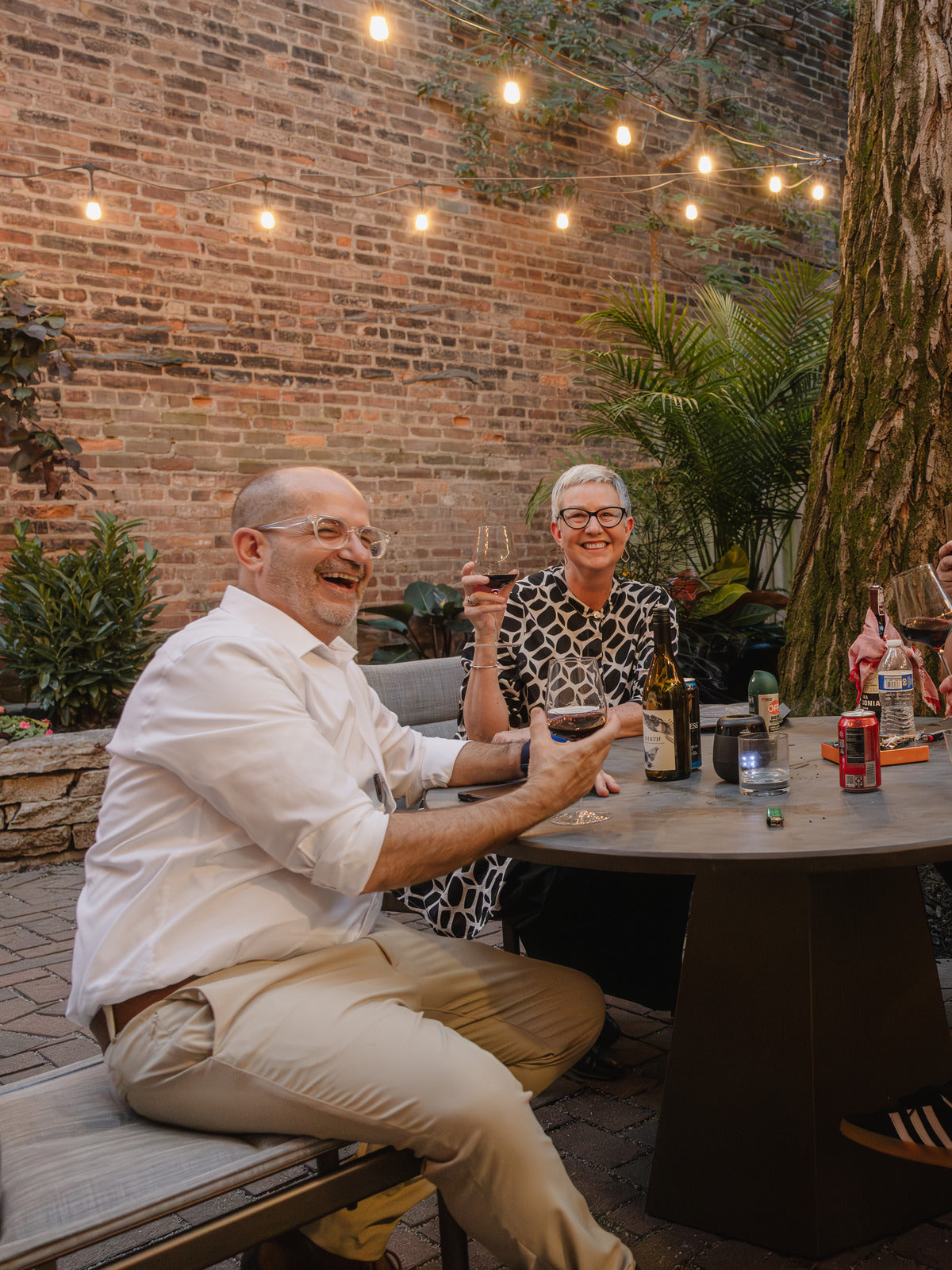 Four friends sharing wine at an outdoor patio table, string lights strung overhead.